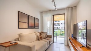 A modern living room with a sofa, a TV, and a balcony with a view of buildings. - Apt w/ Balcony, BBQ & Pool View Ano0209 (Cabo Frio)