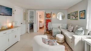 Ein modernes Wohnzimmer mit einem weißen Sofa, einem Couchtisch und einem Fernseher auf einem Standfuß. Eine Tür führt in einen anderen Raum, und an der Wand hängt ein Spiegel.