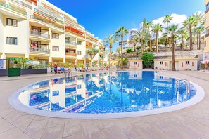 Una piscina rodeada de edificios de varios pisos con balcones, palmeras y un cielo despejado.