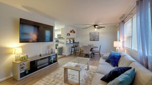 En moderne stue med et fladskærms-tv, et glasbord, en beige sofa med blå puder, et spiseområde med bord og stole, og et køkken i baggrunden.