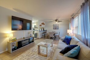 A modern living room with a flat-screen TV, a glass coffee table, a beige sofa with blue cushions, a dining area with a table and chairs, and a kitchen in the background. - U-Med District Townhome w/ Private Yard + Grill (Anchorage)