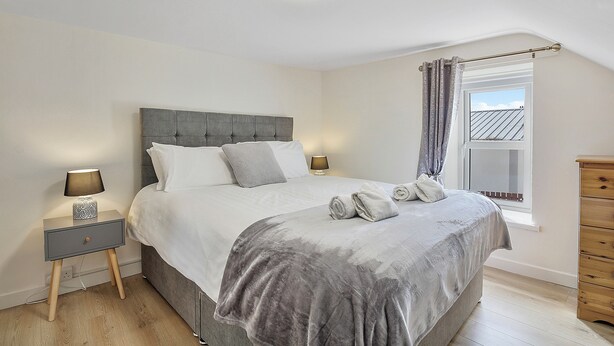 A neatly made bed with a grey headboard, a bedside table with a lamp, and a window with curtains. - The Wee House - Fáilte Ireland Quality Assured (Muff)