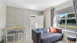 A room with a grey sofa, a wooden dining table with chairs, a window with curtains, and a radiator. - The Wee House - Fáilte Ireland Quality Assured (Muff)
