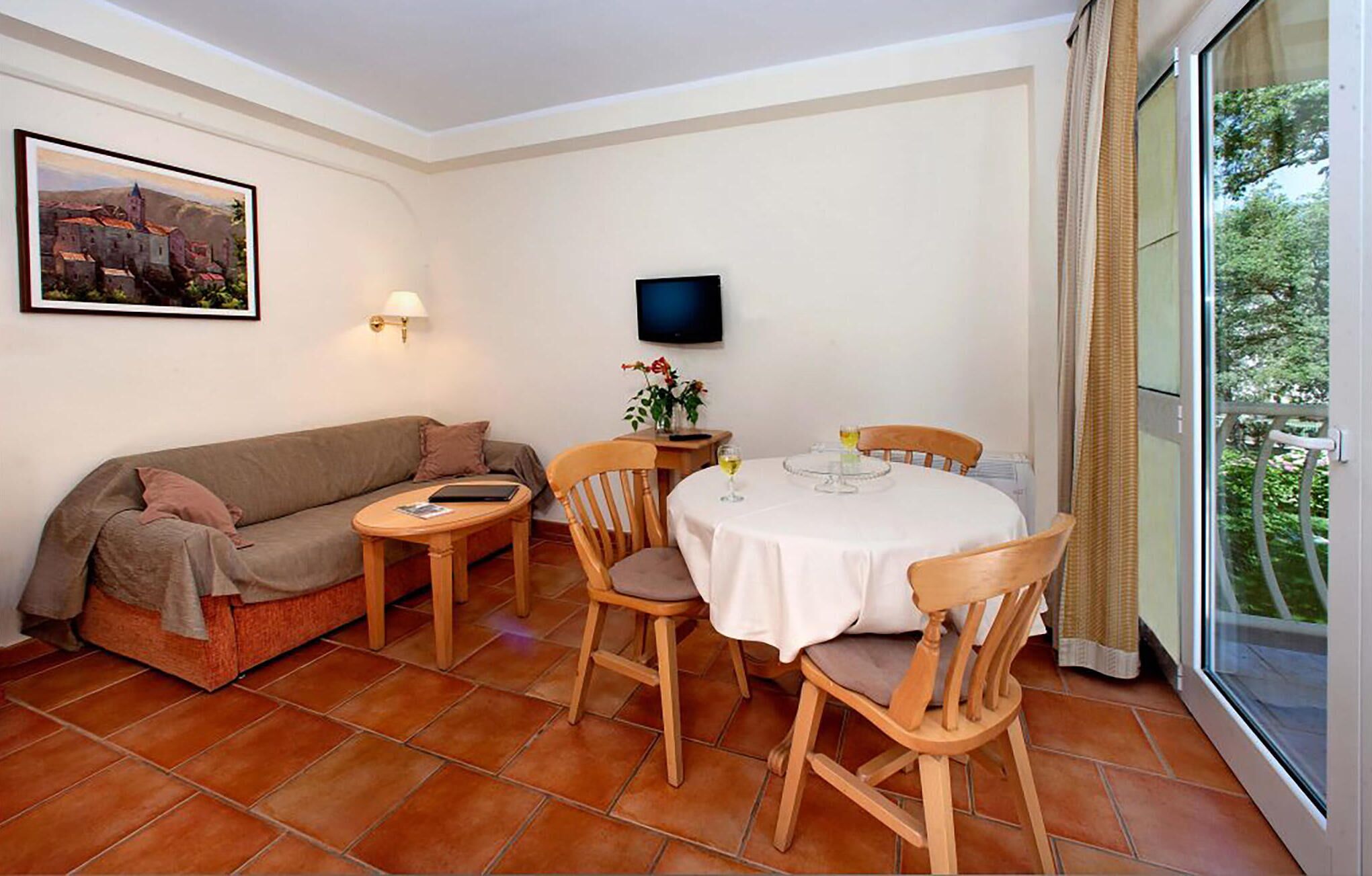A cozy living room with a sofa, a round dining table, and chairs. There's a TV mounted on the wall and a window with a view of greenery.