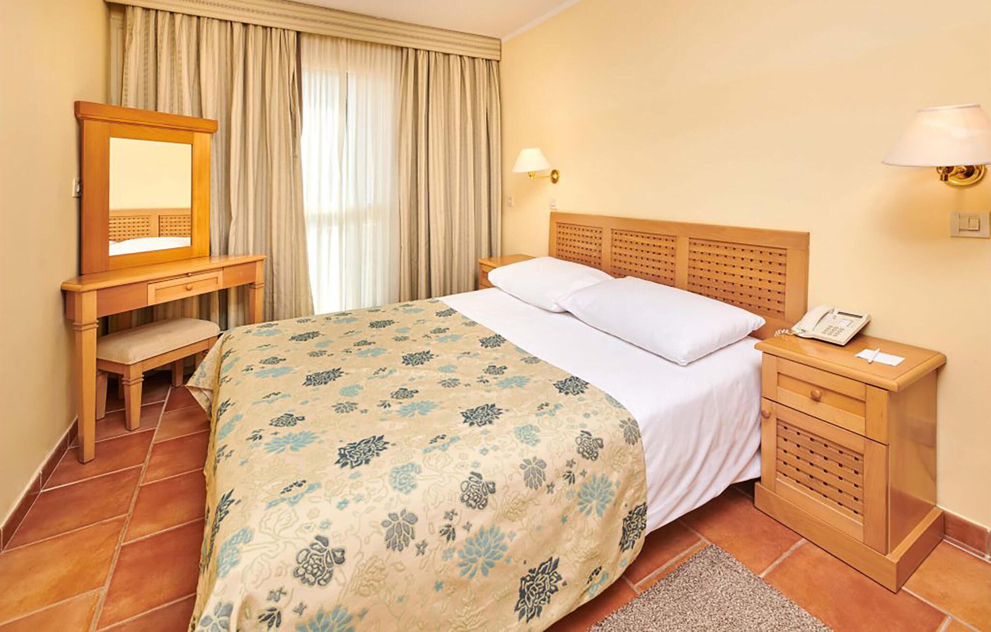 A hotel room with a bed, a desk, a chair, a mirror, a bedside table with a lamp, and a telephone.