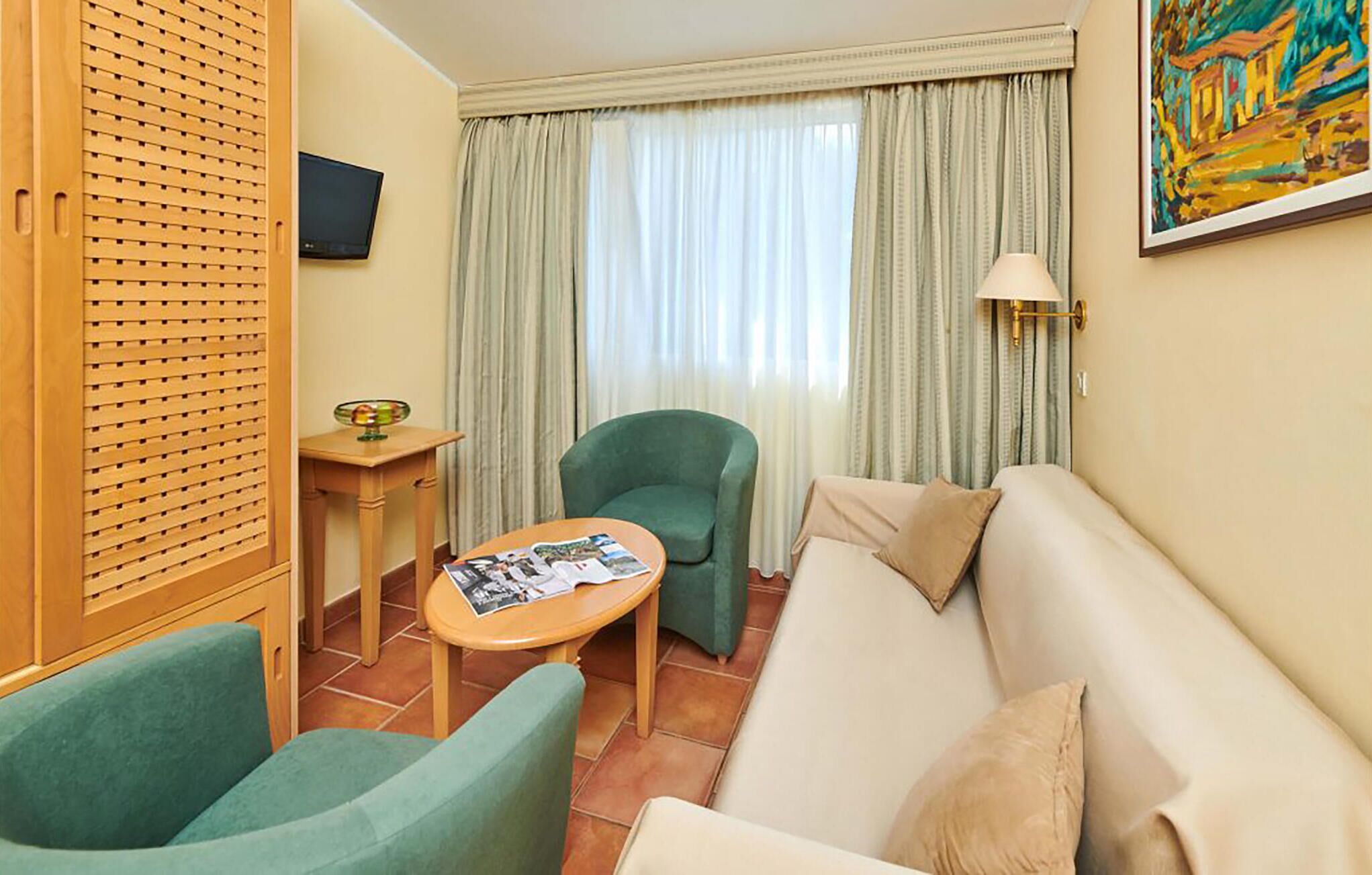 A hotel room with a green armchair, a beige sofa, a small table with magazines, a TV on the wall, and a window with curtains.