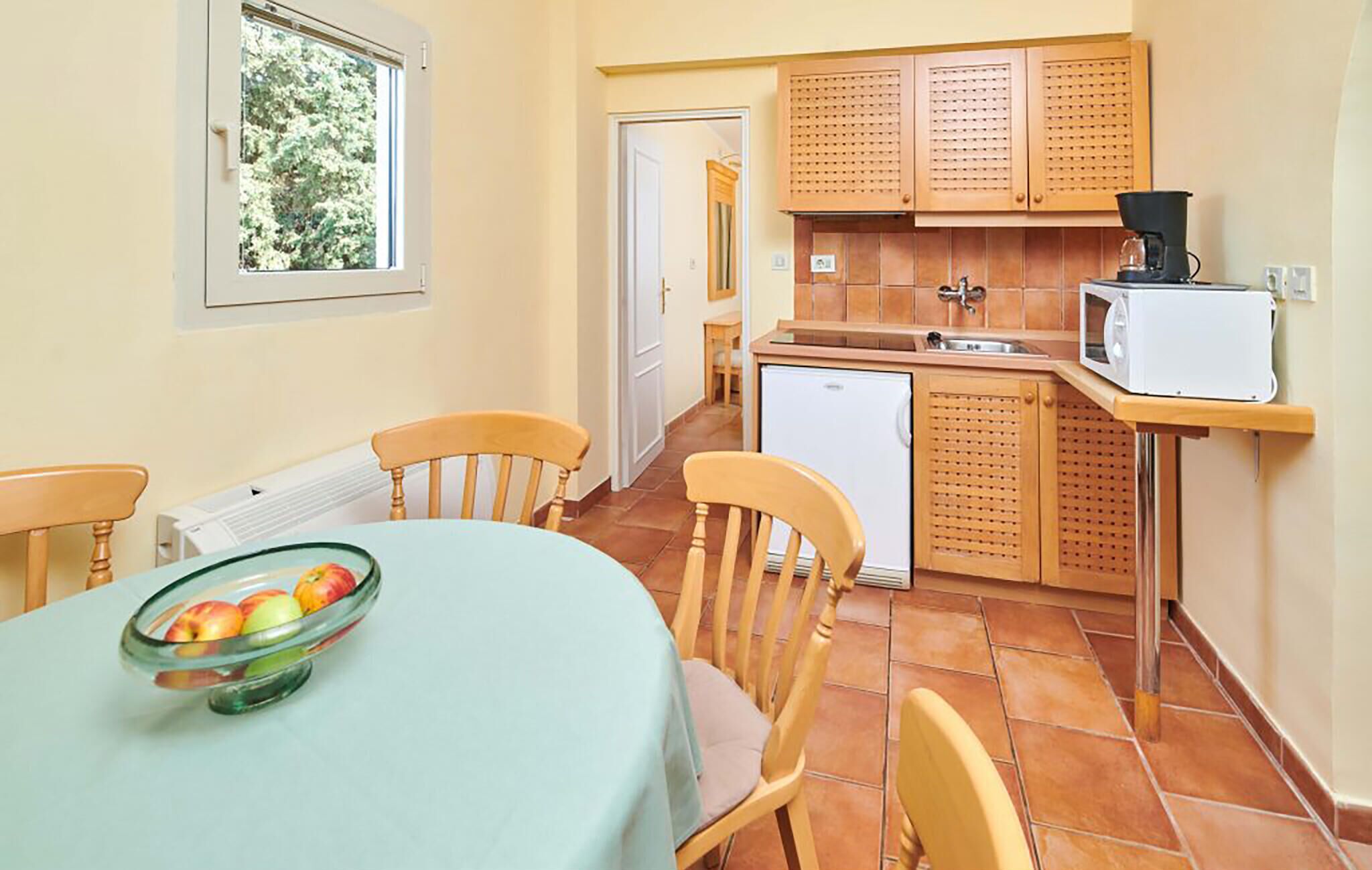 A compact kitchen with wooden cabinets, a white microwave, and a small refrigerator. There is a dining table with a glass bowl of fruit and two chairs.