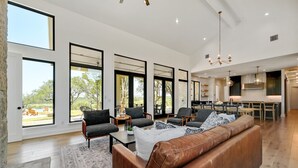 A spacious living area with a large brown leather sofa, multiple armchairs, and a dining table. Large windows provide ample natural light and views of the outdoors. - Ledgestone Ranch - Luxury Ranch - Pickleball - Bocce - Pool - Firepit (Wimberley)
