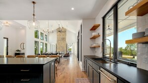 A modern kitchen with a central island, black cabinetry, and a large window providing a view of the outdoors. - Ledgestone Ranch - Luxury Ranch - Pickleball - Bocce - Pool - Firepit (Wimberley)