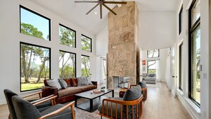 A spacious living room with a stone fireplace, large windows, and a modern ceiling fan. - Ledgestone Ranch - Luxury Ranch - Pickleball - Bocce - Pool - Firepit (Wimberley)