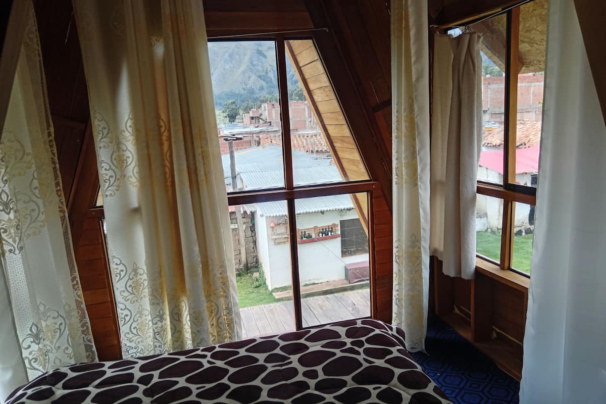 A room with a bed, a window with curtains, and a view of buildings and mountains.