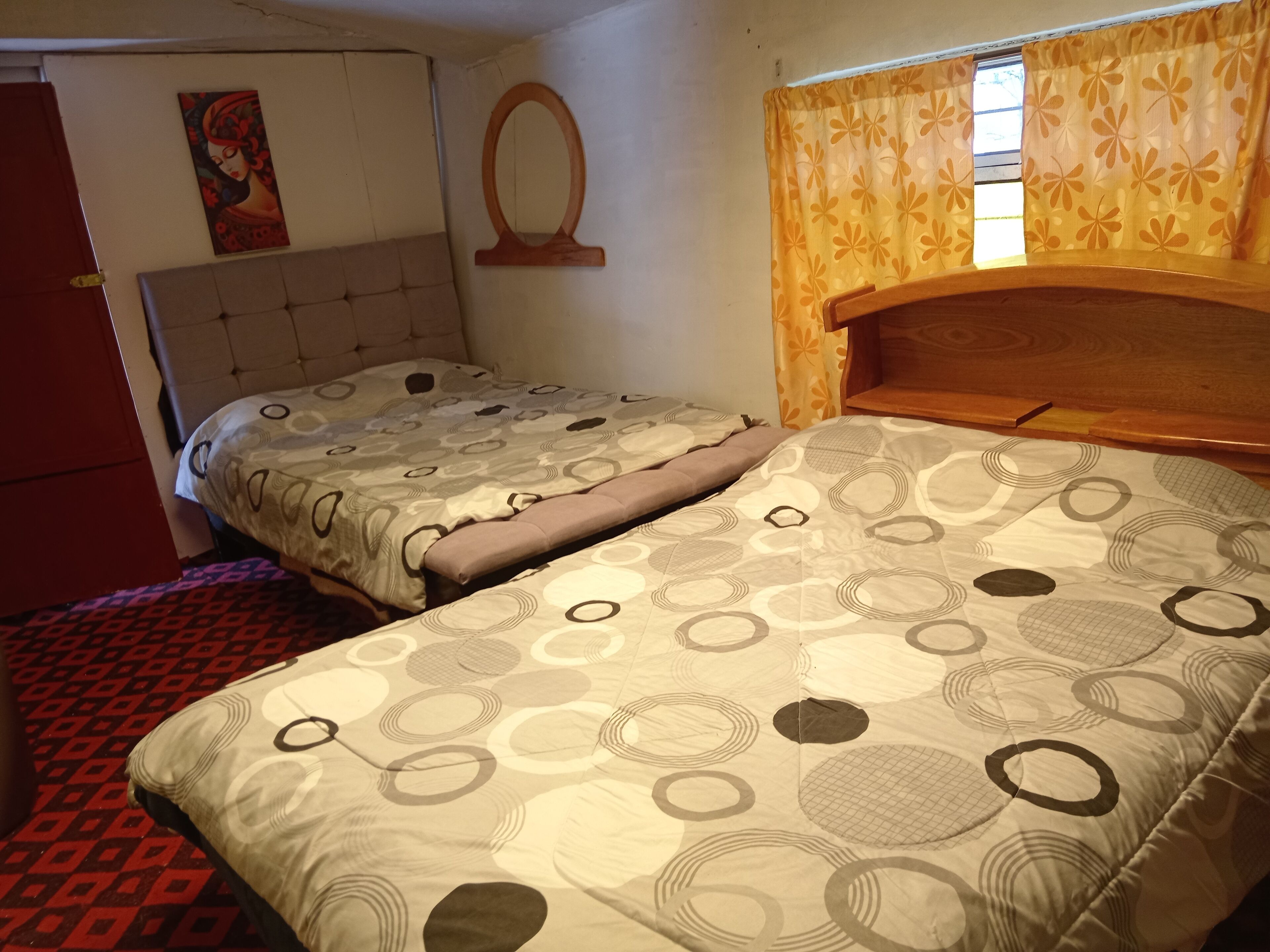 A room with two beds, a red door, a wall painting, a wooden headboard, and a window with curtains.