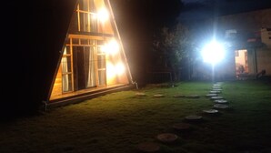 A triangular A-frame structure with a well-lit interior and a grassy yard with stepping stones. - Casa de campo y chalet las morenas  (Sicuani)