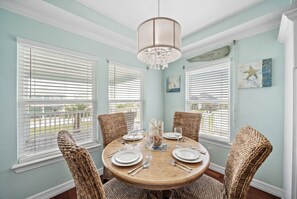 A dining room with a round table, wicker chairs, a chandelier, and a view through the windows.