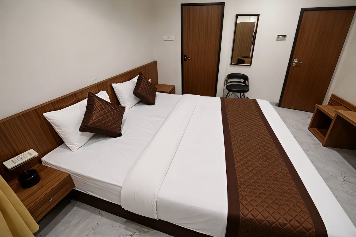 Una cama bien hecha con ropa de cama blanca y marrón, un cabecero de madera y una mesita de noche con una lámpara. Se ven dos puertas y un espejo en la pared.