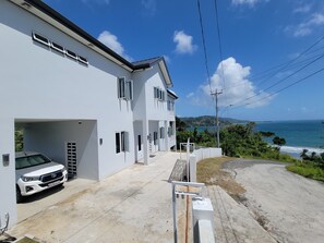 A white two-story house with a car parked in the driveway, overlooking a coastal view. - Speranza - Modern oceanview living in Hope Estate (Hope Estate)