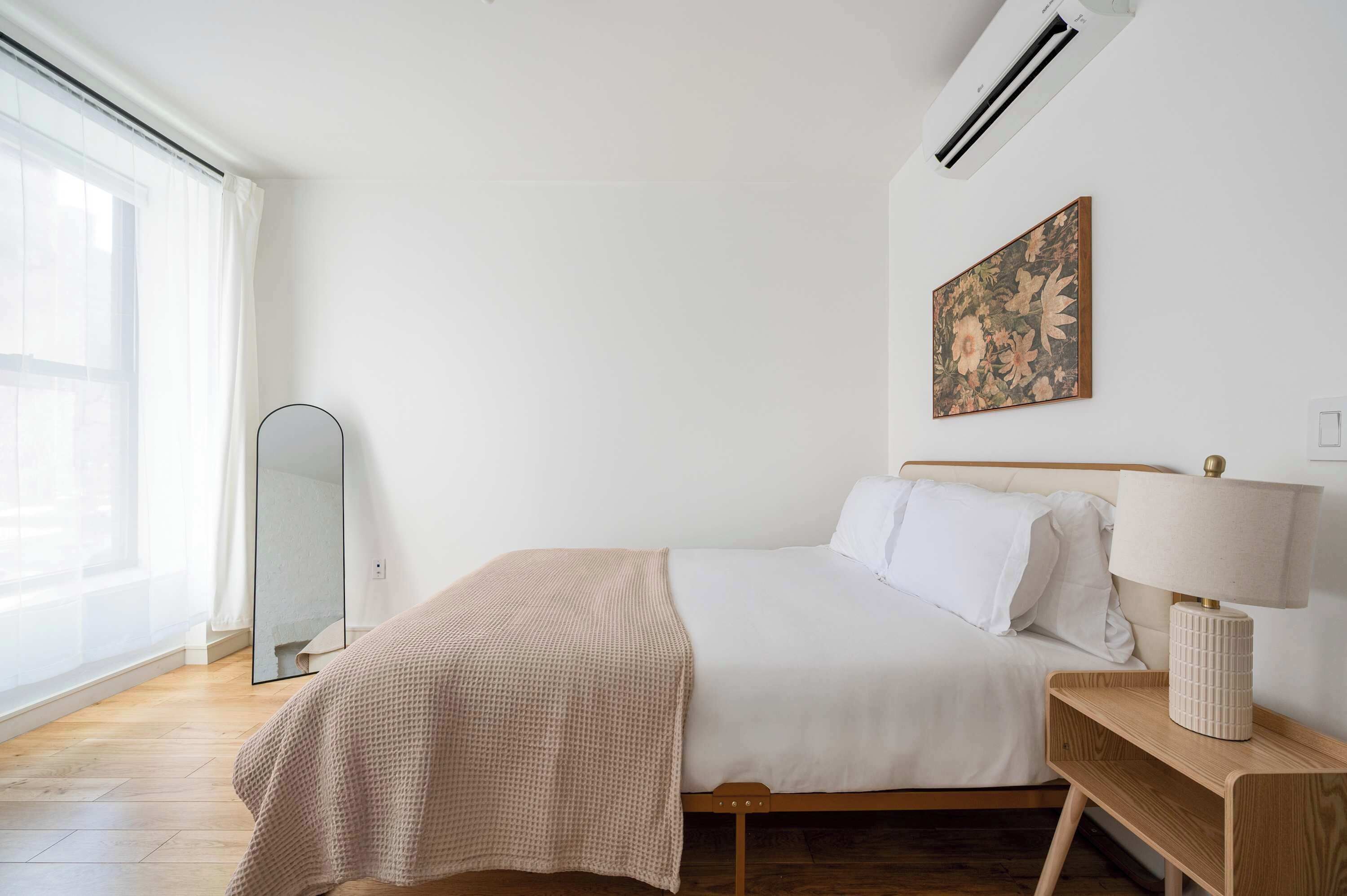 A bedroom with a bed, a bedside table with a lamp, a wall-mounted air conditioner, and a large window with sheer curtains.