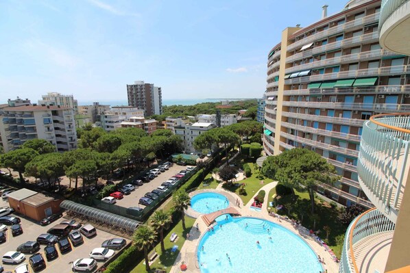A high-rise hotel with multiple balconies, a swimming pool, and a parking area. - Holiday apartment mit Pool und Balkon (Lignano Sabbiadoro)