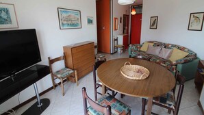 A living room with a TV, wooden furniture, a round table, and a sofa. - Holiday apartment mit Pool und Balkon (Lignano Sabbiadoro)