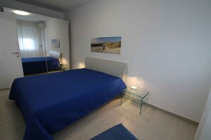 Een slaapkamer met een bed, een glazen bijzettafeltje, een lamp, een foto aan de muur en een spiegel.