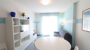 En moderne stue med et rundt hvidt bord, en grå sofa og et hvidt reolsystem med dekorative genstande.