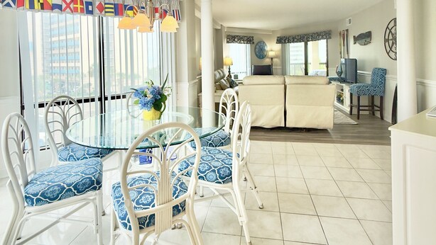 Un comedor con una mesa de vidrio y sillas blancas, una cocina con encimera blanca y una silla azul, y una sala de estar con sofá y televisor.