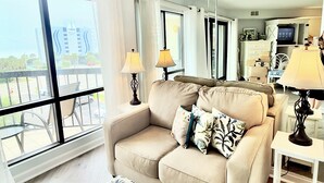 Una sala de estar moderna con un sofá color beige, un reposapiés estampado y una mesita auxiliar blanca. Un espejo grande refleja la habitación y un balcón con mobiliario exterior.