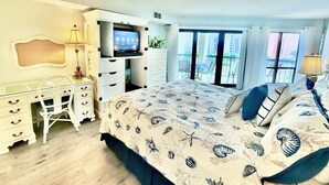 Un dormitorio con cama, escritorio, televisor y balcón con vistas a edificios.