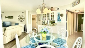 Un comedor con una mesa redonda de vidrio, sillas blancas con cojines de estampado azul, una araña y una cocina al fondo.