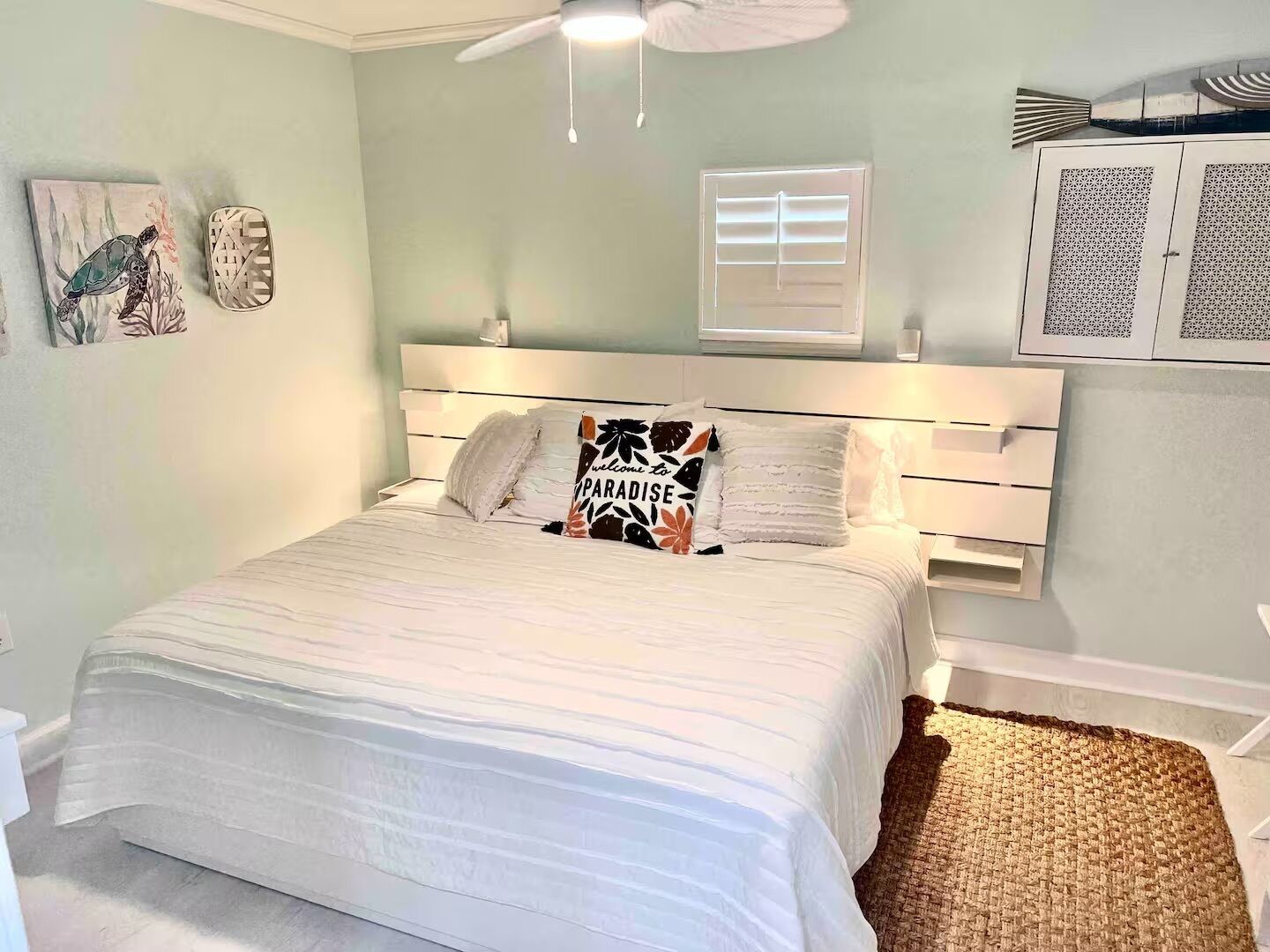 A bedroom with a bed, a ceiling fan, a window with blinds, and a wall-mounted cabinet.
