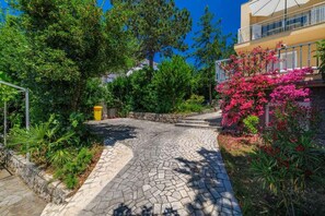 A paved pathway leading to a building with a balcony, surrounded by lush greenery and flowering plants. - Holiday apartment with air conditioning and balcony (Selce)