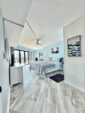 A modern bedroom with a large bed, a ceiling fan, and a view of the city through large windows. - 3BR/3BA Relax, Rewind, Enjoy, Oceanview & Coastal Breeze (Myrtle Beach)