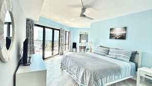 A modern bedroom with a large bed, a desk, a chair, and a view of the city through large windows. - 3BR/3BA Relax, Rewind, Enjoy, Oceanview & Coastal Breeze (Myrtle Beach)