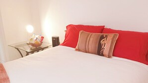 A neatly made bed with red and beige pillows, a glass table with a fruit basket, and a bedside lamp.