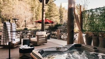 A hot tub on a wooden deck with outdoor furniture, a red umbrella, and a forest backdrop.