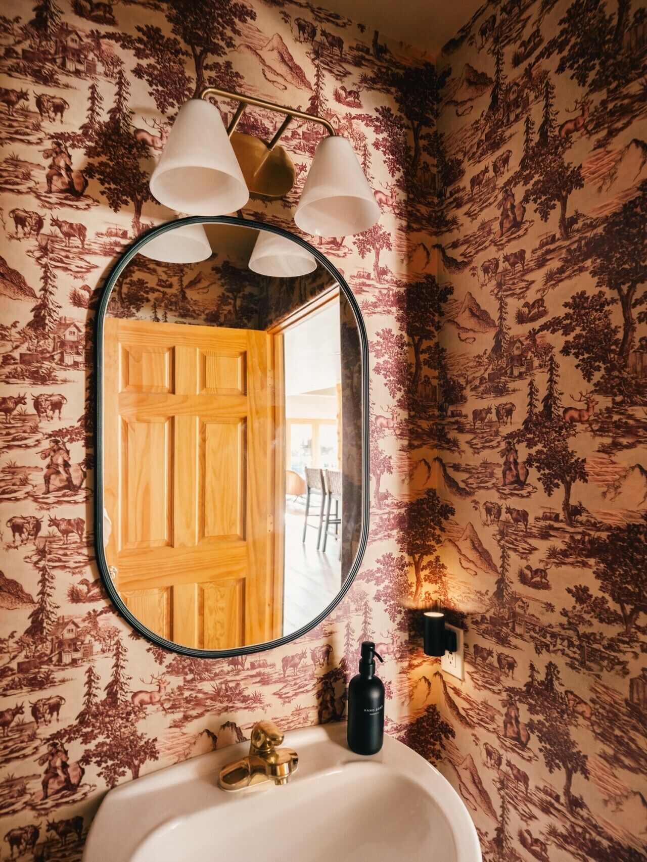 Une salle de bain avec une porte en bois, un miroir rond, un lavabo et un mur recouvert d’un papier peint à motifs naturels.