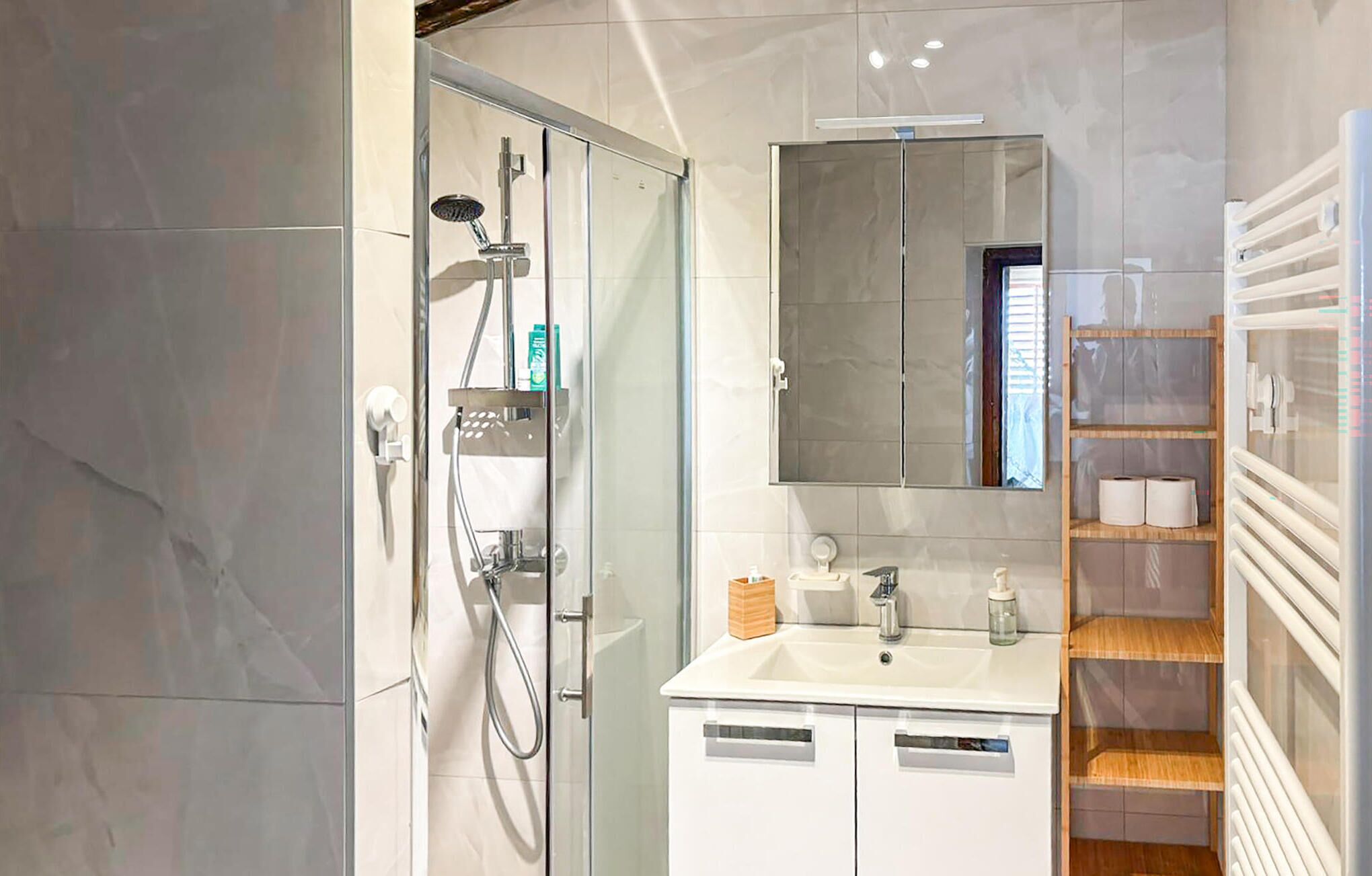Un baño moderno con cabina de ducha de vidrio, un lavabo blanco, un espejo y un estante de madera con artículos de aseo.
