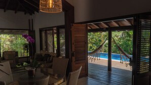 A tropical resort room with a glass table, wicker furniture, and a view of a pool and hammock. - Luxury Nature Retreat: Hummingbird House (Tibau do Sul)