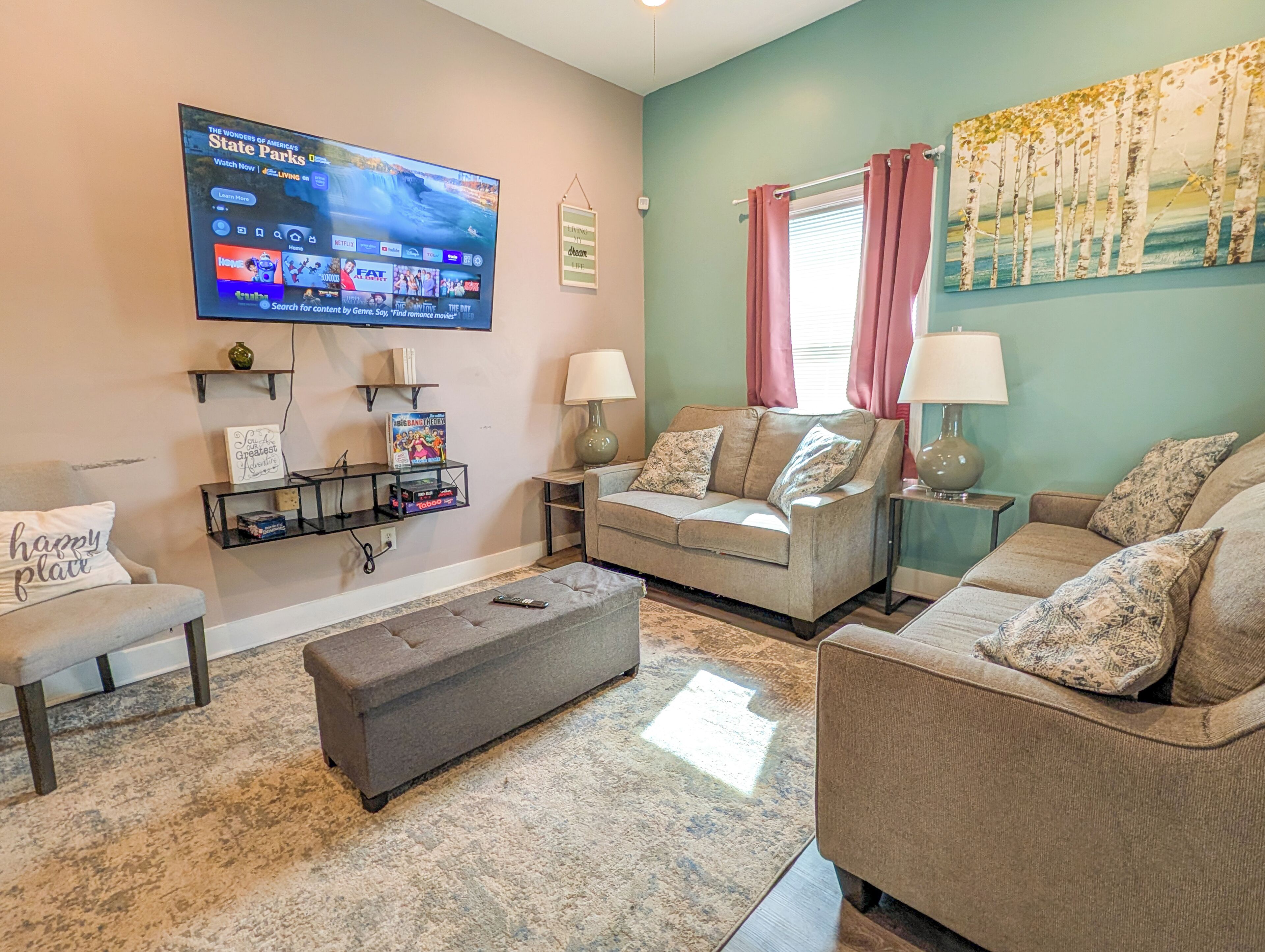 A living room with a flat-screen TV, two sofas, a coffee table, a side table, a lamp, a painting, and a shelf with books and a vase.