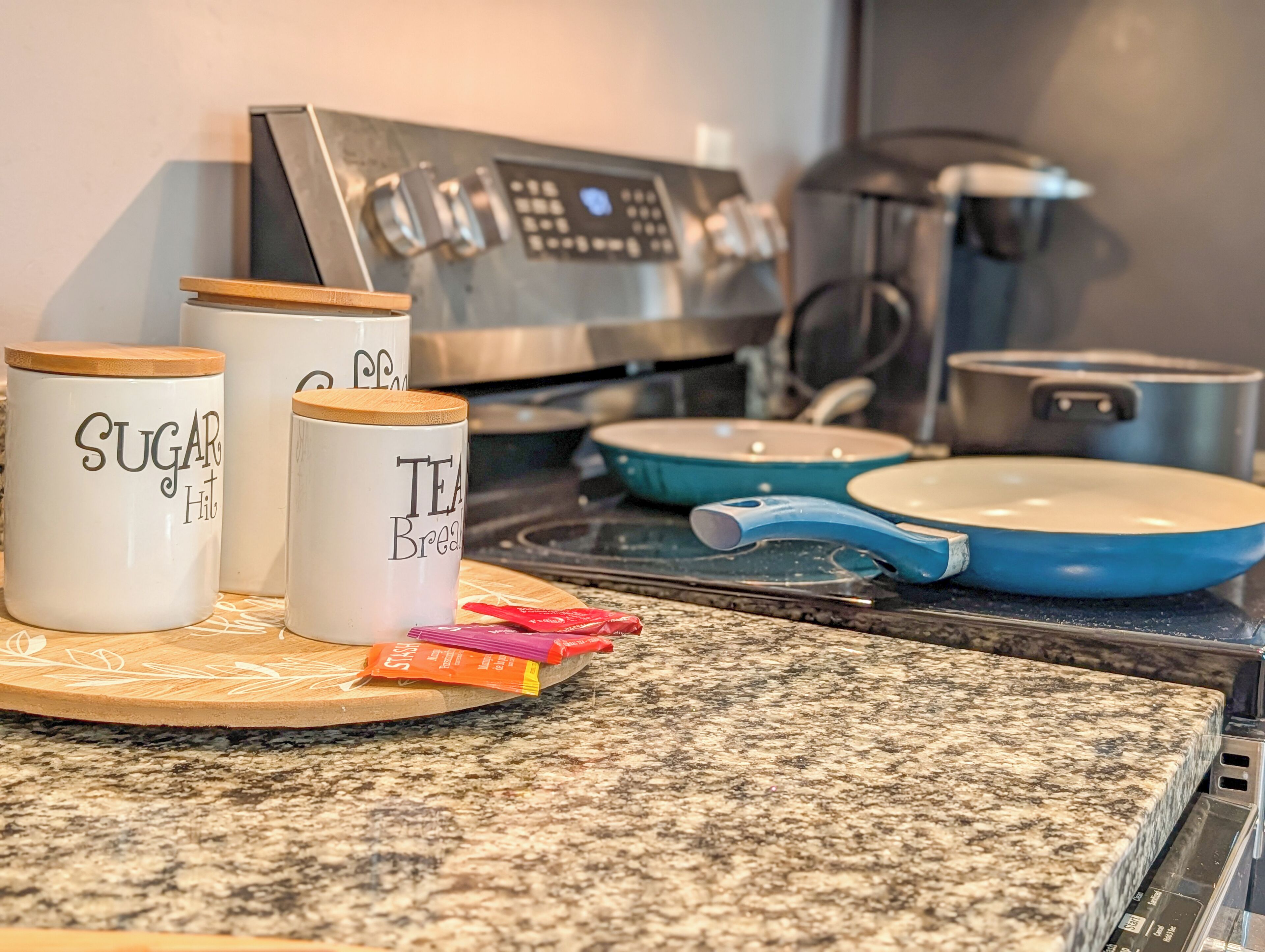Et køkken med en kaffemaskine, to krukker med påskrifterne 'SUGAR HIT' og 'TEA BREAK', samt en blå pande på bordet.