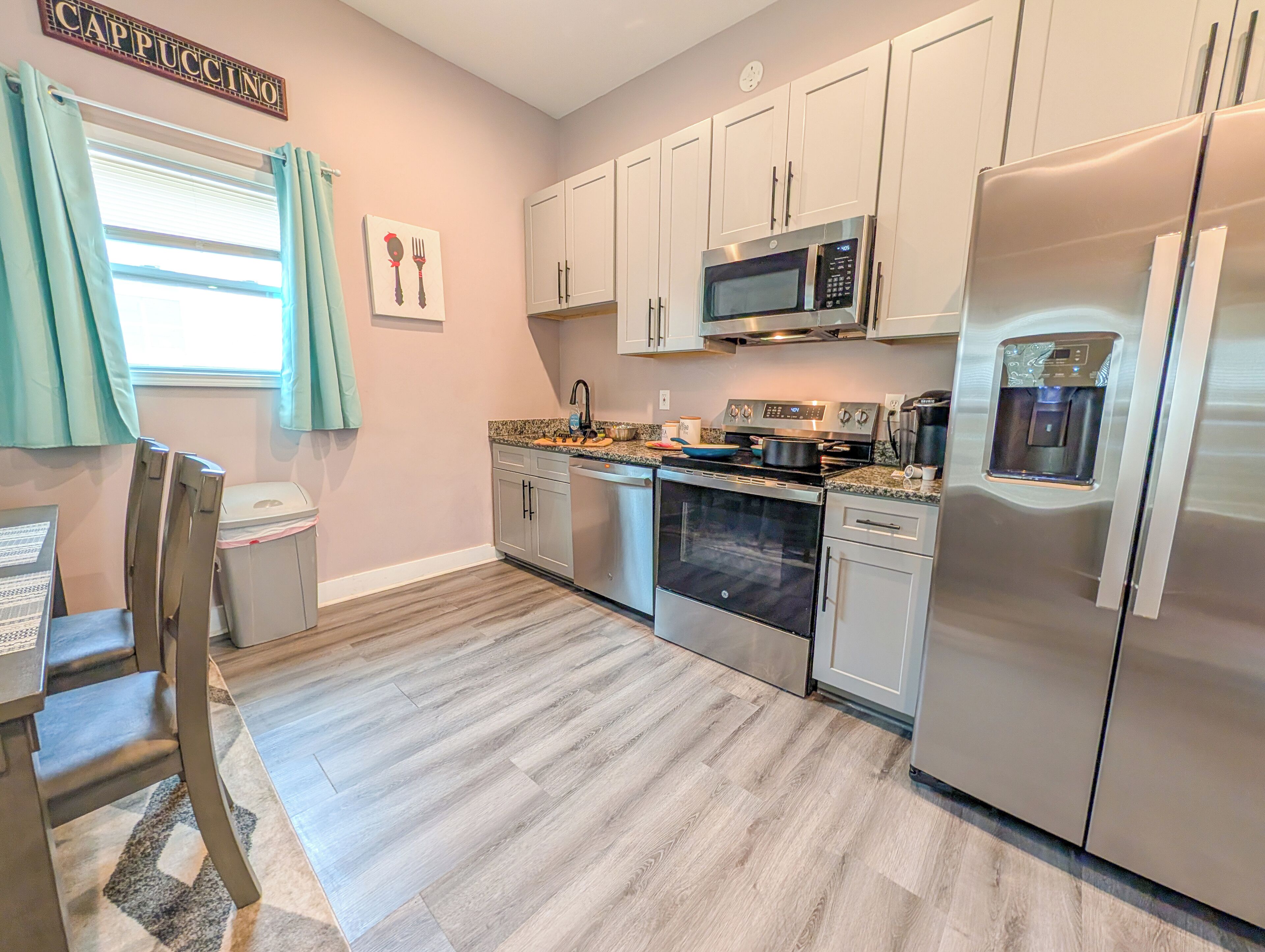 A modern kitchen with stainless steel appliances, a microwave, oven, and a refrigerator. There is a window with curtains, a sink, and a countertop with a coffee maker.