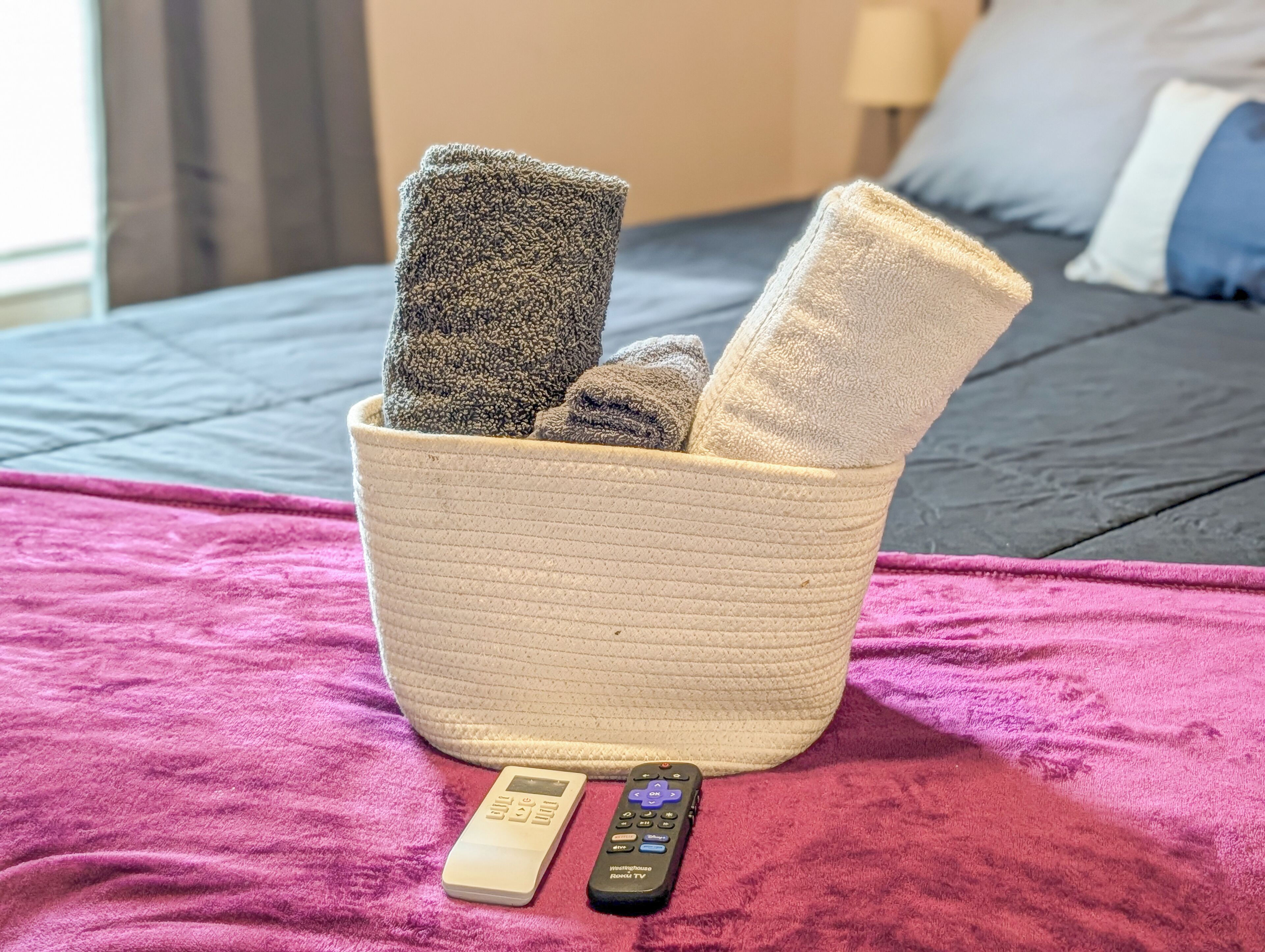 A bed with a pink blanket, a woven basket with towels, and two remote controls on the bed.