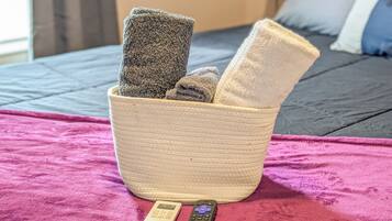 A bed with a pink blanket, a woven basket with towels, and two remote controls on the bed.