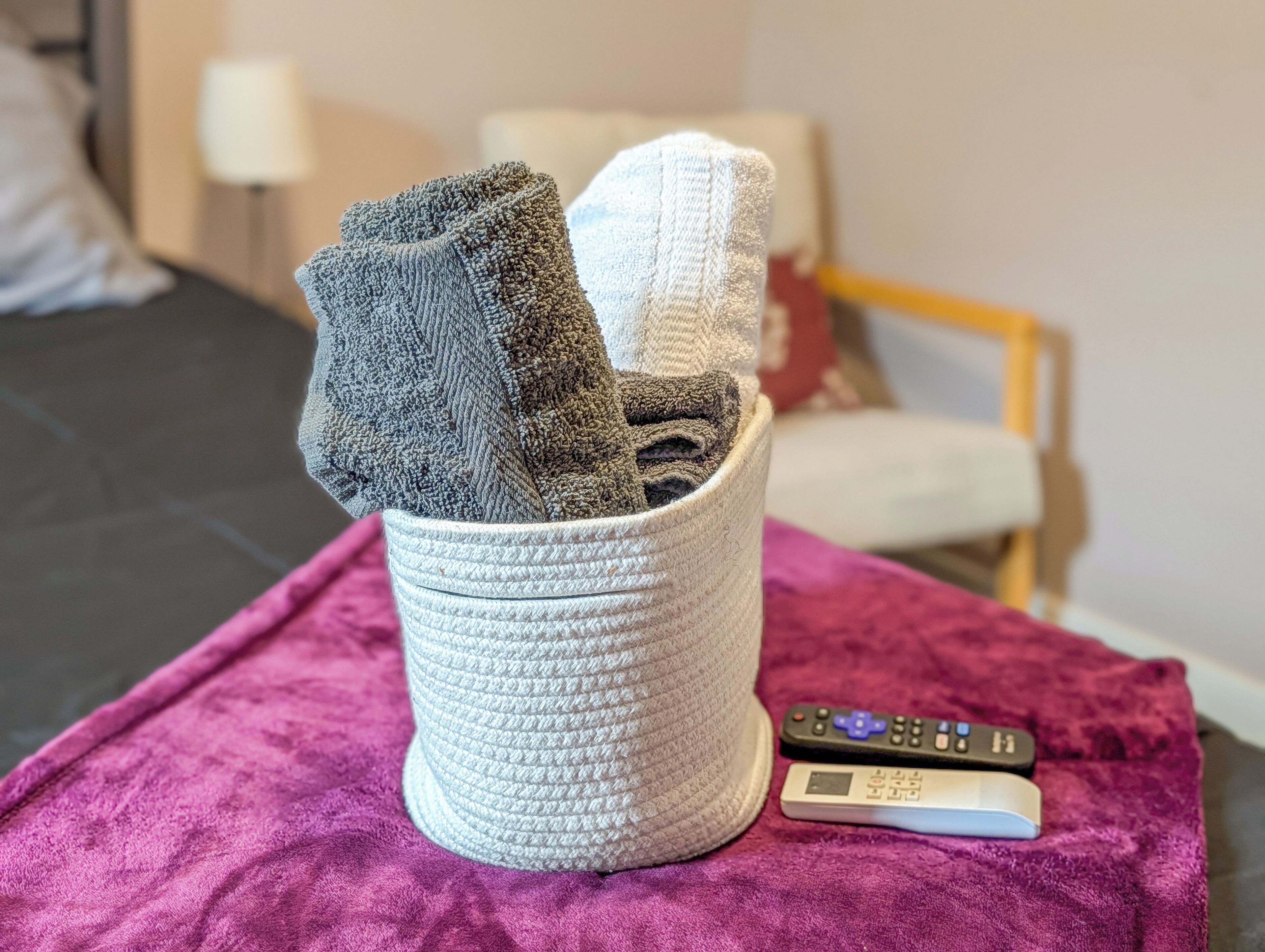 Een bed met een paarse deken, een witte cilindrische container met opgevouwen handdoeken, en een afstandsbediening op het bed.