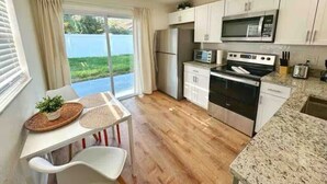 A modern kitchen with white cabinets, a stainless steel refrigerator, a microwave, and an oven. There is a dining table with chairs and a view of a green landscape through the window. - NEW Gorgeous Sunshine-Up 6 Guests-Private Backyard (Port Orange)