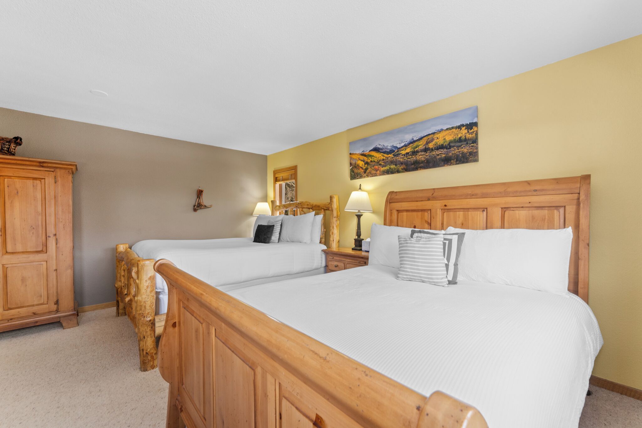 A hotel room with two wooden beds, a wooden wardrobe, a nightstand with lamps, and a painting on the wall.