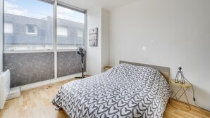 A bedroom with a large bed, a standing fan, a small table with a lamp, and a window with a view of buildings.