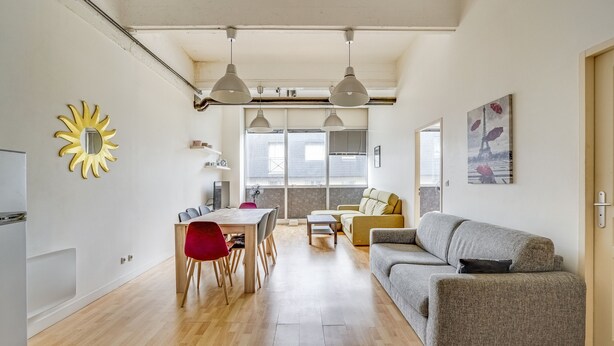 A modern living space with a dining area, a sofa, and a large window providing natural light.
