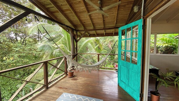 A wooden deck with a hammock, a turquoise door, and a view of lush greenery.