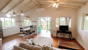 A spacious living area with a sofa, a dining table, and a TV. There is a kitchen area with a stove and a sink. The room has wooden floors and a ceiling fan.